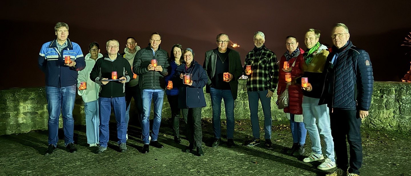 In Dunkelheit steht ein Gruppe von 12 Personen draußen. Sie halten kleine Leuchter in der Hand.