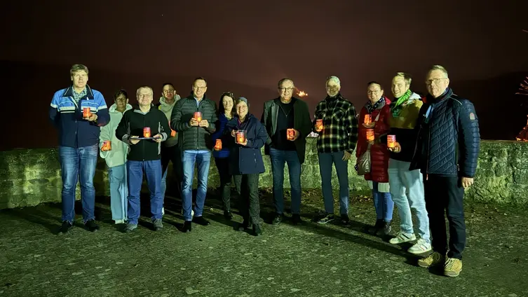 In Dunkelheit steht ein Gruppe von 12 Personen draußen. Sie halten kleine Leuchter in der Hand.