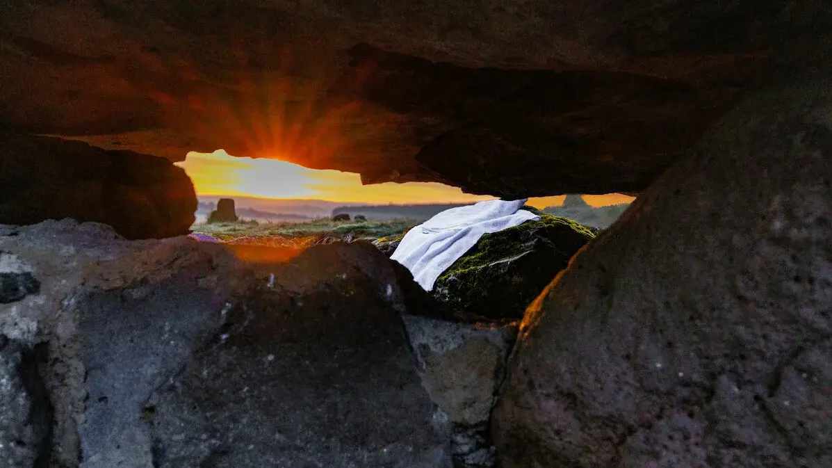Man sieht das Leichentuch von Jesus vor der Höle liegen mit Blick aus der Höle. Im Hintergrund sieht man die Sonne aufgehen.