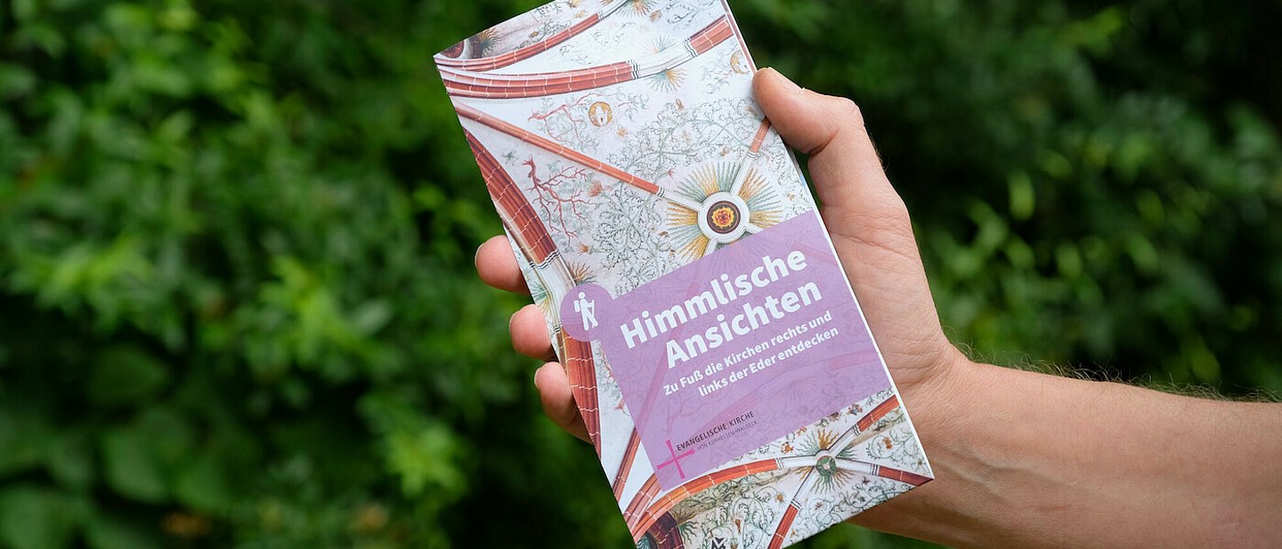 Eine weiße Hand hält die bunt gemusterte Broschüre / Wanderkarte mit der Aufschrift "HImmlische ANsichten" in die Kamera.