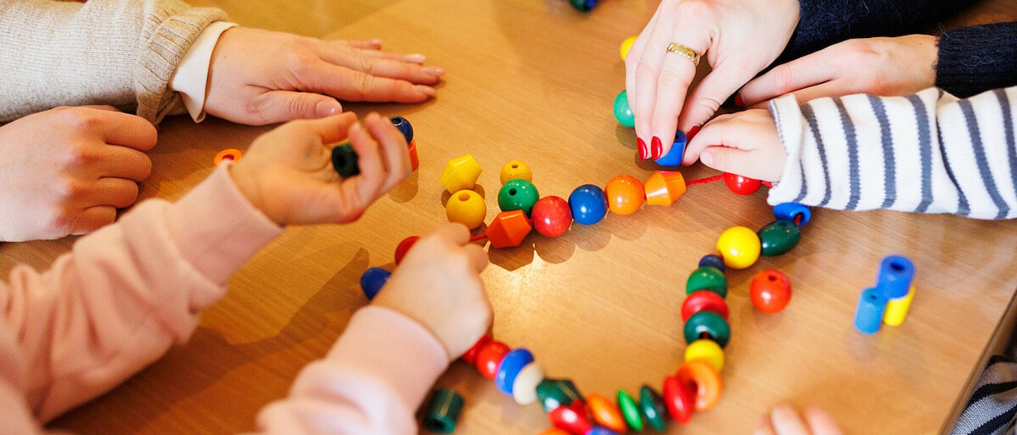 Kita - Kinder beim Spielen. An einem Tisch sitzen einige Kinder, eine Erzieherin und spielen mit einer Kette mit bunten Kugeln und anderen Formen, die auf einer Schnur aufgereiht sind.