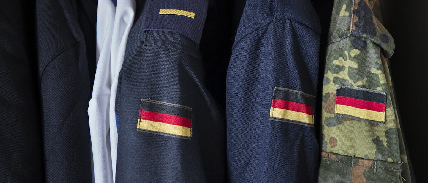 Bundeswehr-Uniformen sind nebeneinander aufgehängt. Auf der linken Schulter ist jeweils eine kleine Deutschlandflagge aufgenäht.