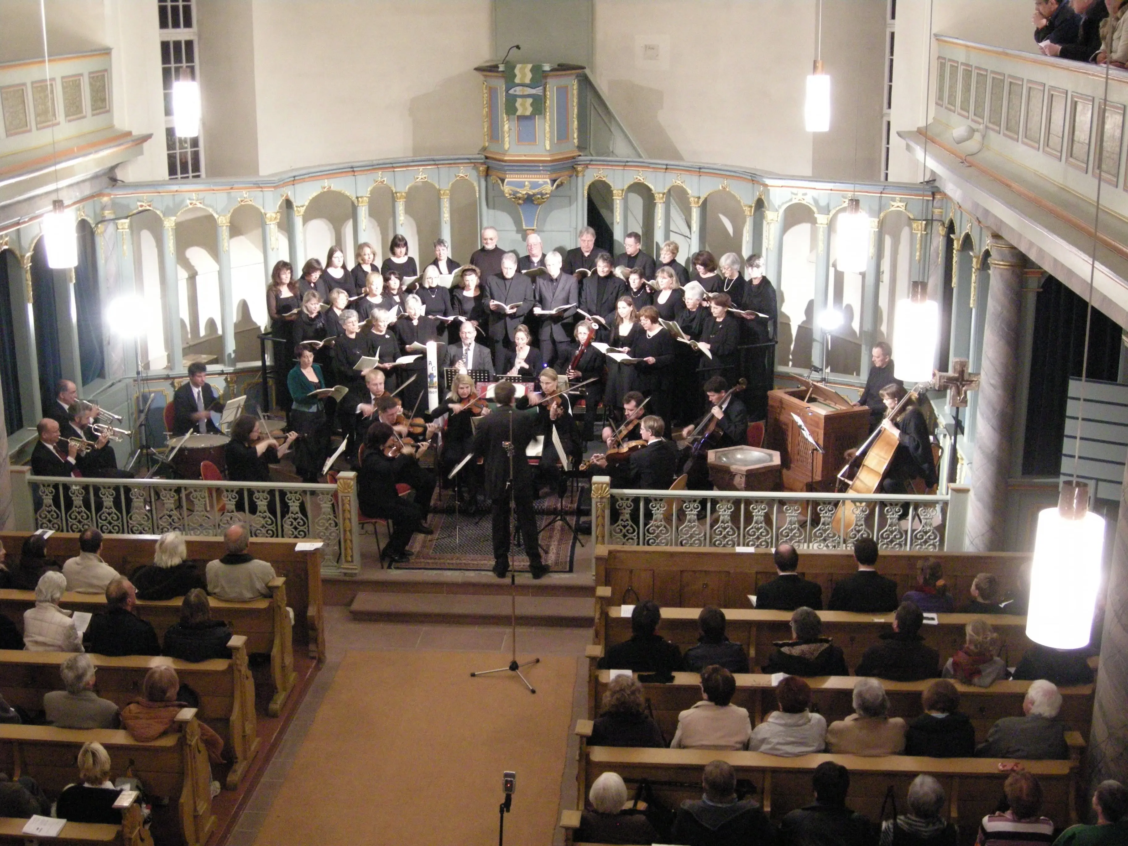 Foto von der Empore einer Kirche auf den Altarraum in dem ein Chor (gekleidet in Anzügen und schwarzer Kleidung) singt. Die Kirchenbänke sind mit Zuschauenden gefüllt. Es singt die Taunussteiner Kantorei