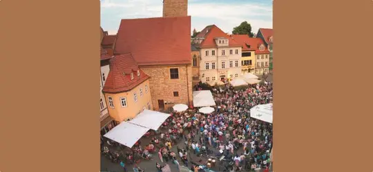 Luftaufnahme eines belebten Platzes in einer Altstadt mit vielen Menschen, Sonnenschirmen und historischen Gebäuden.