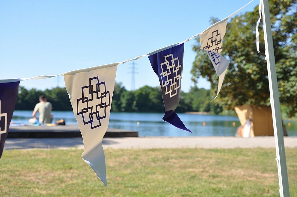 Zu sehen sind Wimpel mit dem Logo der EKHN beim Tauffest in Bensheim. Im Hintergrund befindet sich ein Badesee.