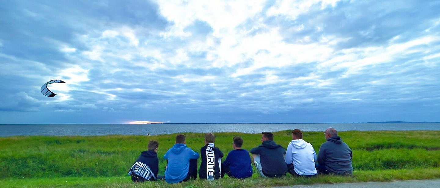Man sieht eine Gruppe Jugendlicher am Strand sitzen die einem Kiter bei Ihrem Sport zusehen. Im Hintergrund sieht man den mit Gras bewachsenen Strand mit dem Meer dahinter. Der Drachen des Kiters fliegt in dem bewölktem Himmel auf der linken Seite des Bildes.