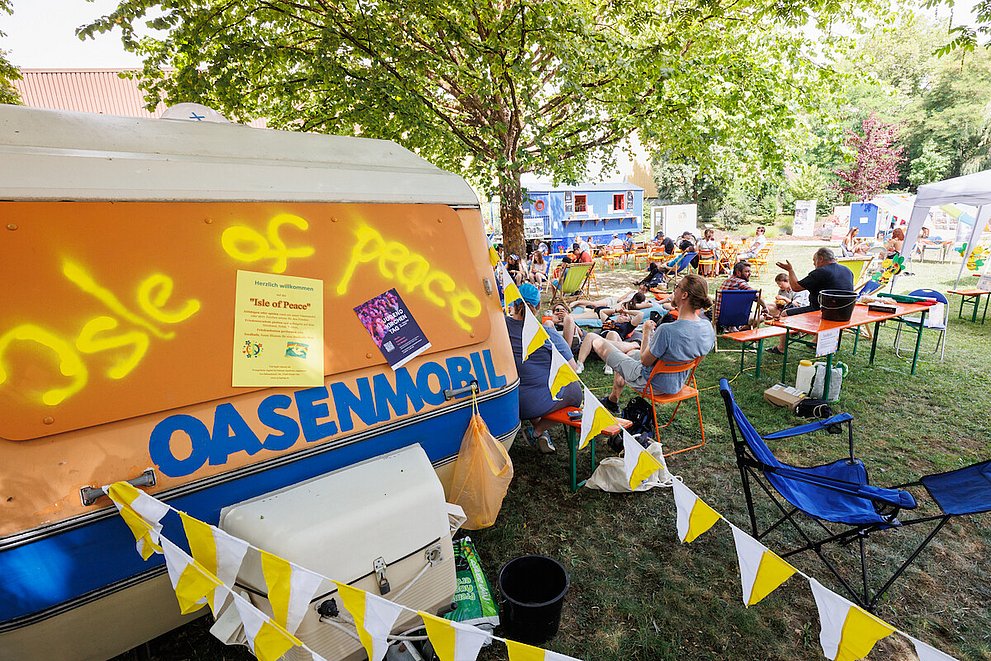 Man sieht im Vordergrund das Oasenmobil und im Hintergrund Jugendliche auf Bänken und Stühlen