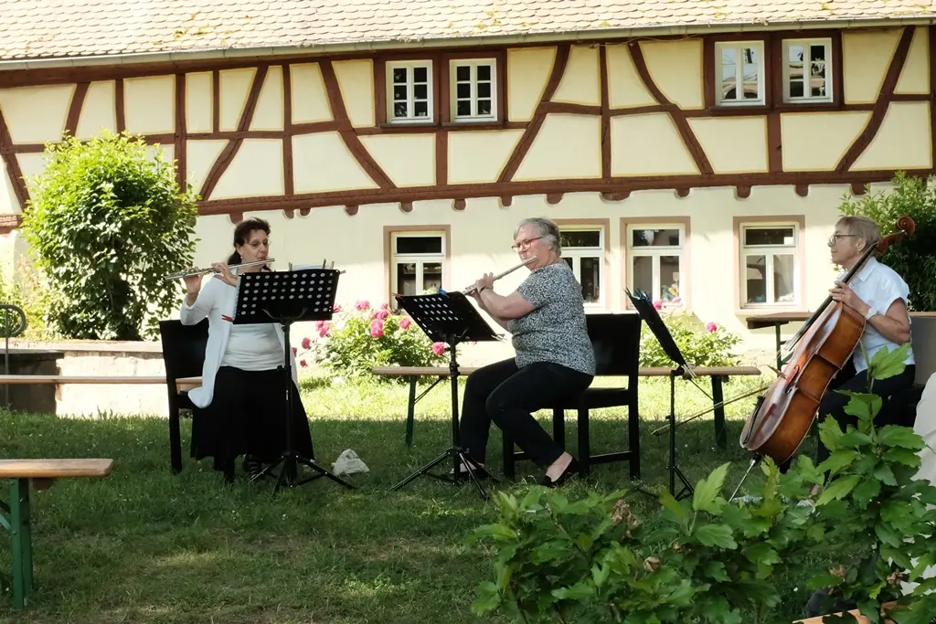 Hier sitzen zwei Querflötistinnen und eine Cellistin des Ensembles in einem Garten eines großen Fachwerkgebäudes und musizieren. 
