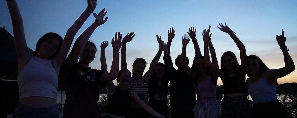 Man sieht eine Gruppe Jugendliche vor dem Abendhimmel, die Gesichter verschattet, die die Arme hoch reißen
