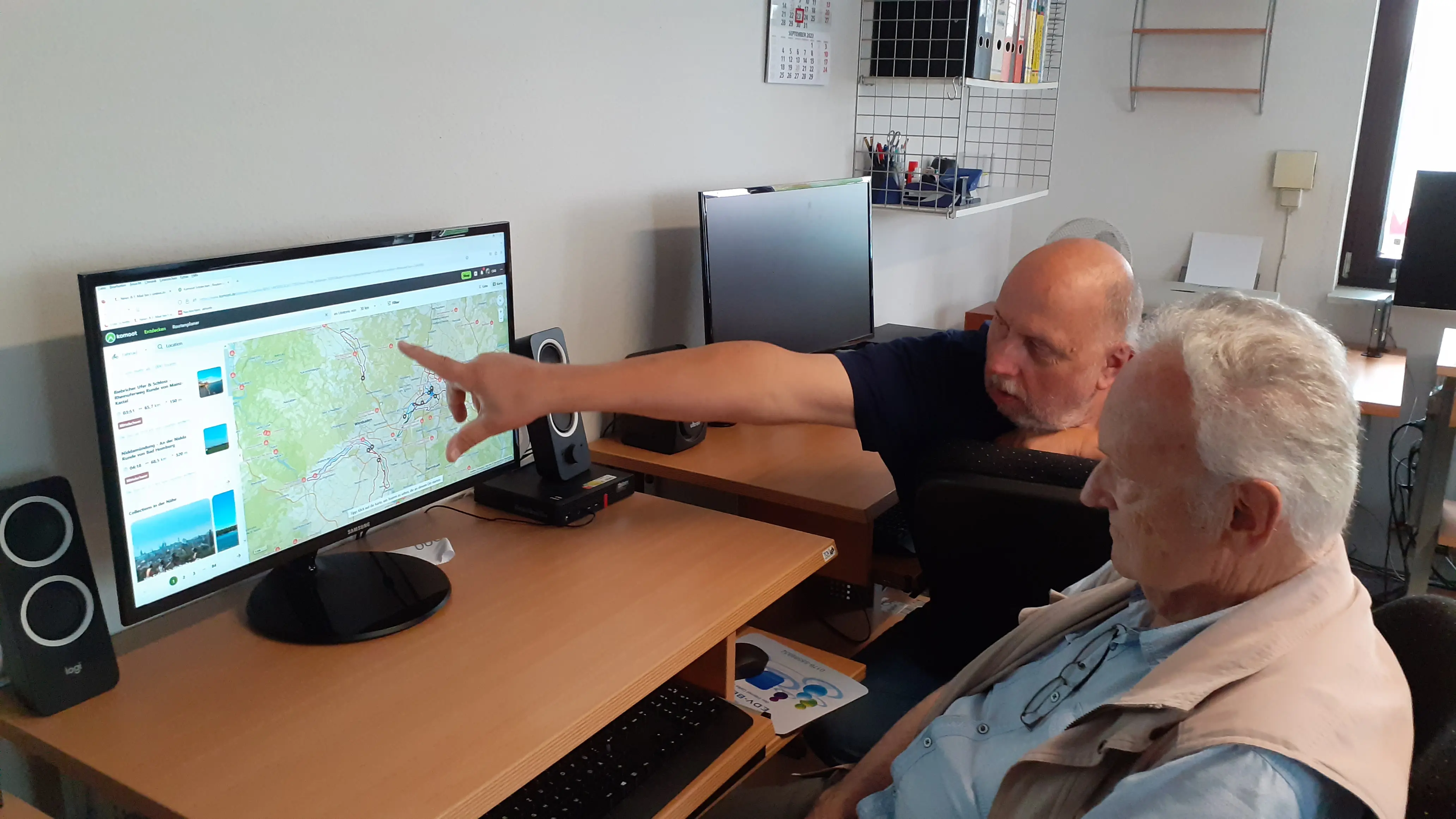 Das Bild zeigt zwei ältere Männer in einem Raum. Einer der Männer, mit kurzem Haar, sitzt an einem Schreibtisch und zeigt auf einen Computermonitor. Der Monitor zeigt eine Karte oder einen Plan. Der zweite Mann, mit weißen Haaren, sitzt vor dem Monitor und schaut ebenfalls darauf. Auf dem Schreibtisch befinden sich Lautsprecher, und im Hintergrund sind andere Bürogegenstände zu sehen.