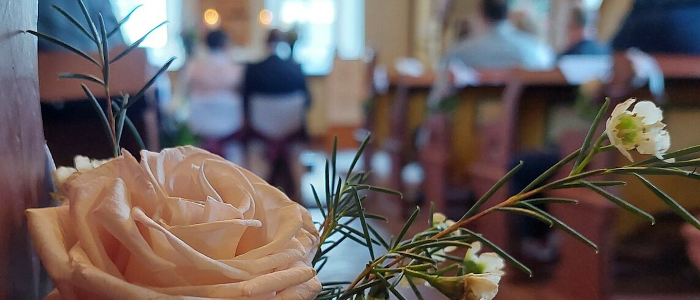 Kirchliche Trauung: Eine helle Rose an einer Kirchenbank steht im Fokus. Im Hintergrund sitzt ein Ehepaar vor dem Altar.