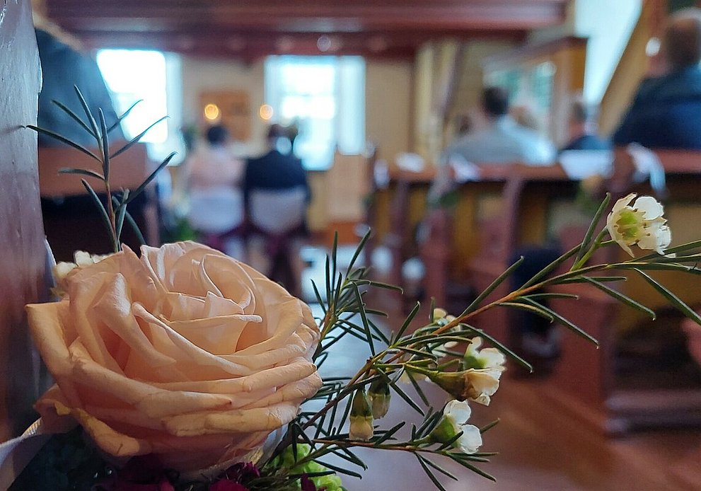 Kirchliche Trauung: Eine helle Rose an einer Kirchenbank steht im Fokus. Im Hintergrund sitzt ein Ehepaar vor dem Altar.
