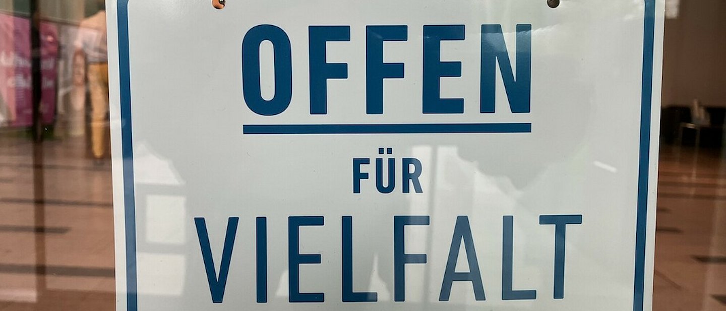 An der Innenseite eines Schaufensters hängt ein Schild mit der Aufschrift "Offen für Vielfalt" in Großbuchstaben. Die Schrift ist blau auf weißem Grund