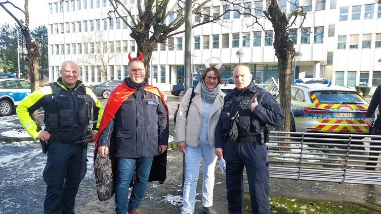 Am Rand stehen Polizeifahrzeuge. Außen stehen zwei Polizeibeamte in Uniform. Der Seelsorger und die Seelsorgerinnen stehen innen und sind als Teufel und Engel verkleidet. 