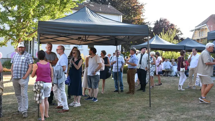 Schlange wartender Menschen im Pfarrgarten, Blick in das Gelände mit Biertischgarnituren, Pavillons und Sonnenschirmen.