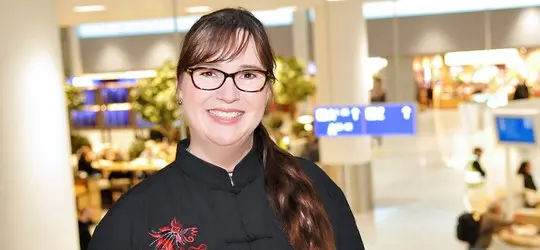 Eine lächelnde Frau mit Brille und langem Zopf steht in einem Flughafengebäude. Sie trägt dunkle Kleidung mit roten Verzierungen, im unscharfen Hintergrund sind Sitzbereiche und Hinweisschilder zu sehen.