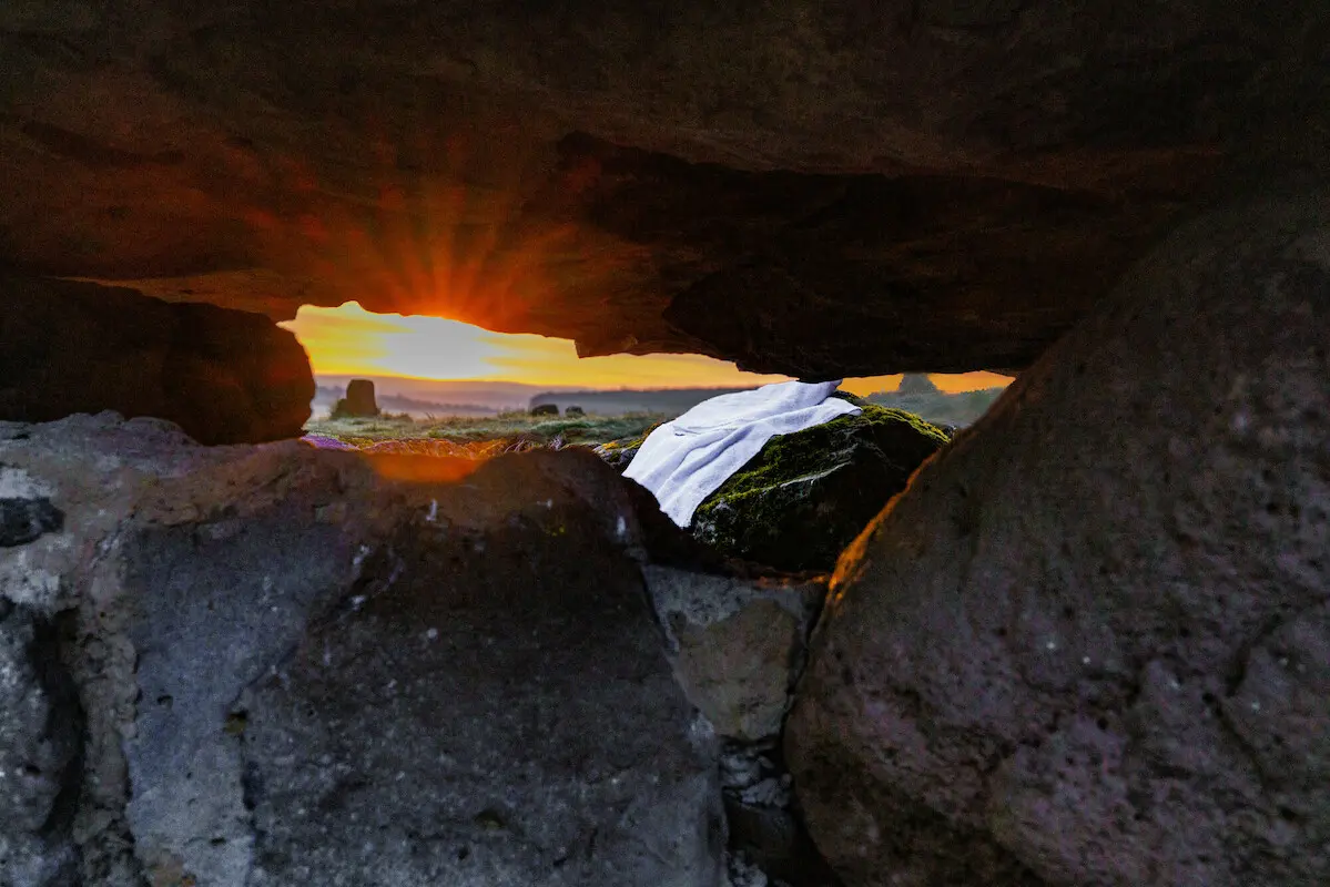 Man sieht das Leichentuch von Jesus vor der Höle liegen mit Blick aus der Höle. Im Hintergrund sieht man die Sonne aufgehen.