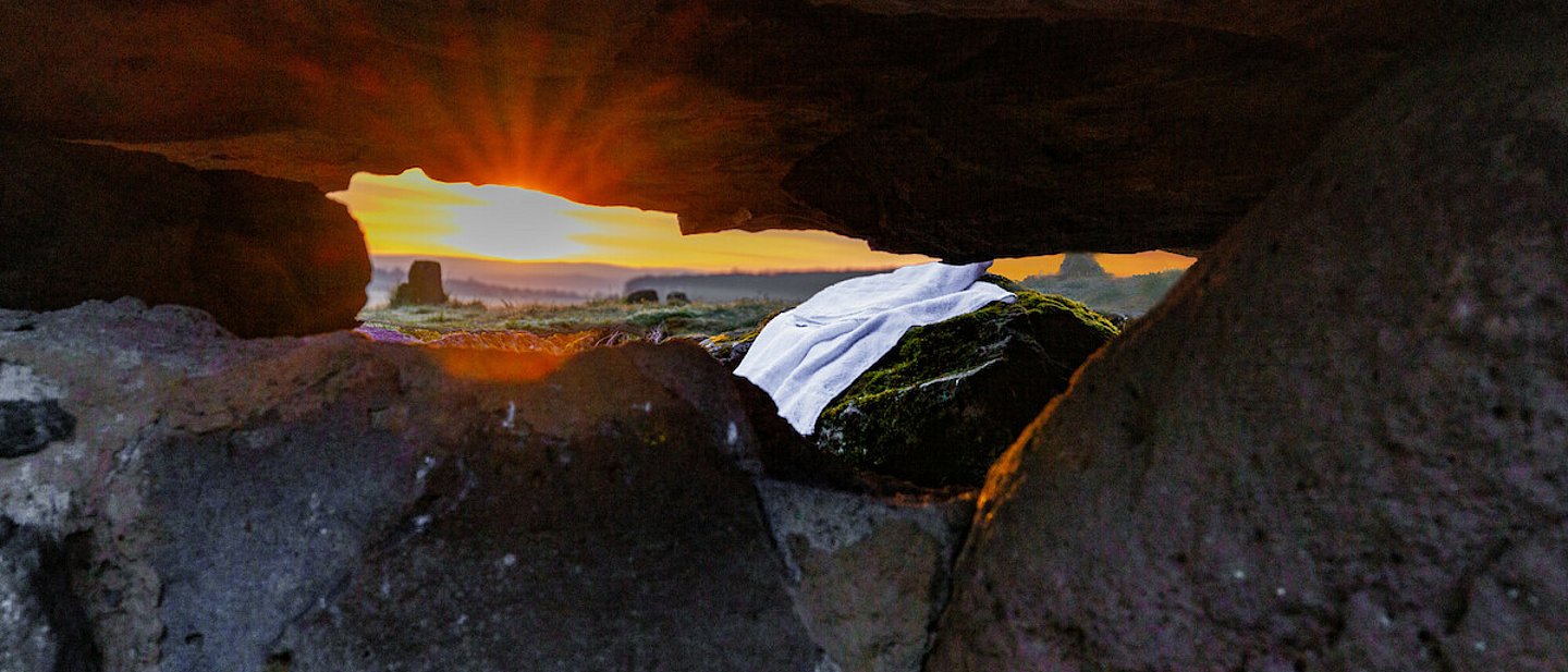 Man sieht das Leichentuch von Jesus vor der Höle liegen mit Blick aus der Höle. Im Hintergrund sieht man die Sonne aufgehen.