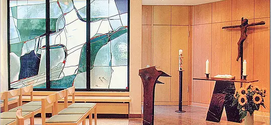 Kleiner Kapellenraum mit Altar, Kruzifix und zwei Kerzen; links stehen Stühle vor einer großen Glasfensterwand mit abstraktem Motiv.