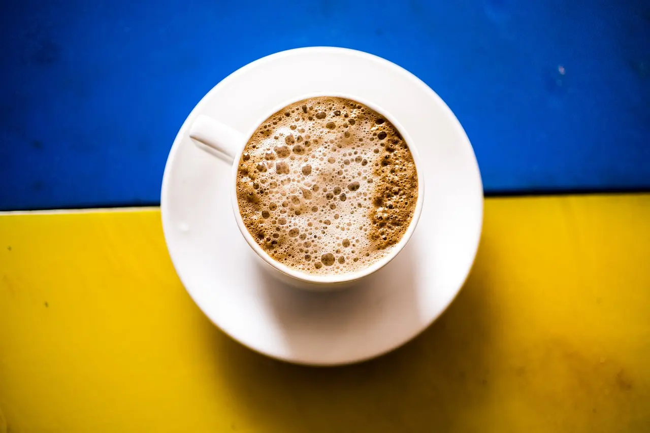 Das ist ein Foto einer Tasse Kaffee auf einem Untersetzer. Die Tasse steht auf einer Fläche, die zur Hälfte blau und zur Hälfte gelb ist – ähnlich den Farben der ukrainischen Flagge. Der Kaffee sieht schaumig aus.