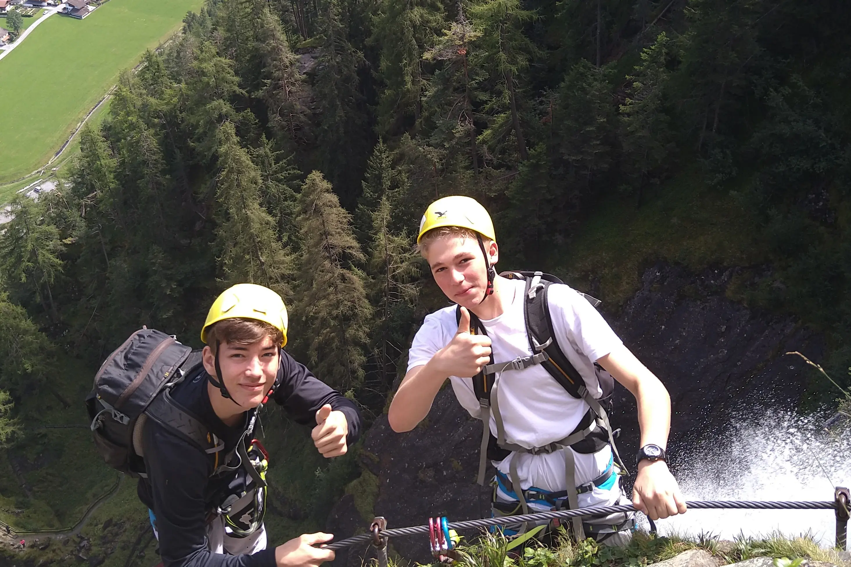 Zwei Jugendliche mit Helm, sind am Gipfel eines Berges angekommen und halten sich am Seil fest und zeigen "Daumen hoch"
