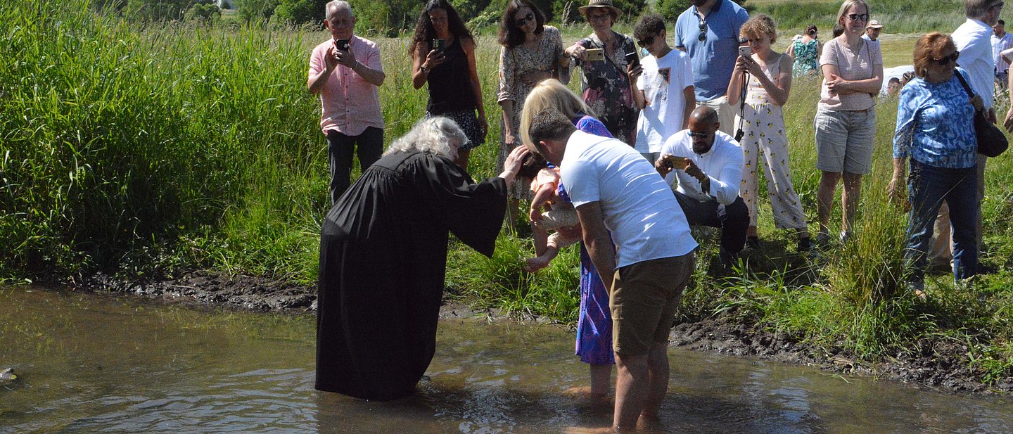 Eine Pfarrerin steht mit Talar im Wasser und tauft ein Kind, das vom Vater gehalten wird. Am Ufer stehen Menschen und schauen zu.
