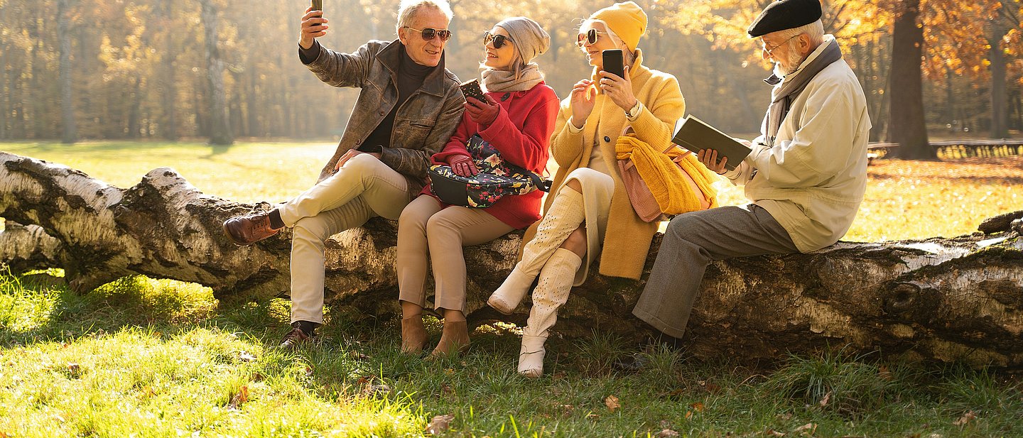 Vier ältere Menschen sitzen vergnügt in der Natur mit Handys und Büchern.