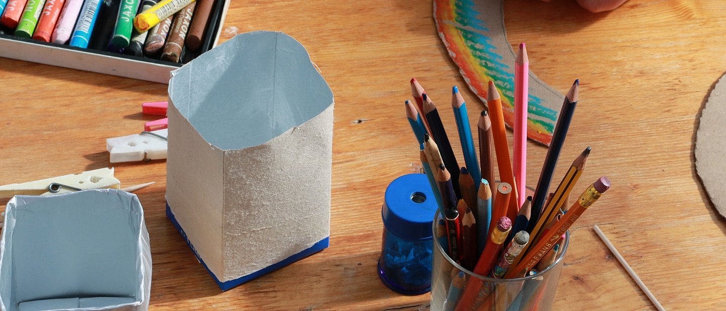 Auf einem Tisch steht ein Becher mit Buntstiften und ein Spitzer, daneben liegen Wachsmalkreiden. Ein abgeschnittener, unbedruckter Tetrapak steht zum Bemalen daneben. Dahinter liegt ein kleiner Regenbogen aus Pappe 
