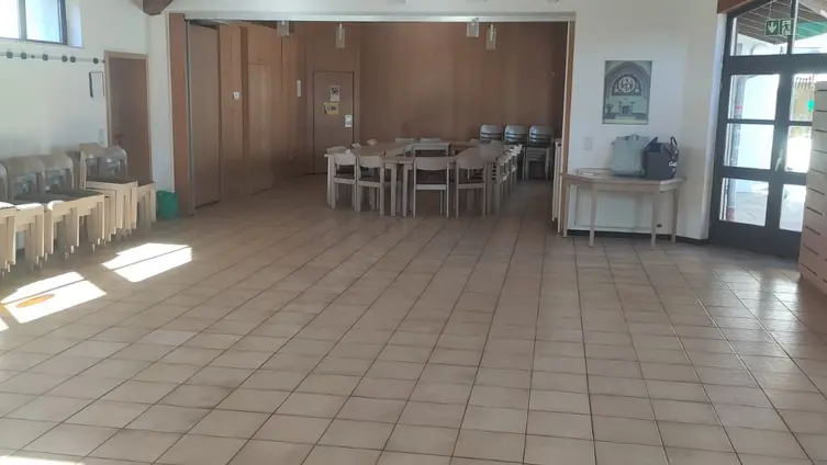 Ein großer, gefließter Raum. Im Hintergrund steht eine Tischgruppe mit Stühlen.