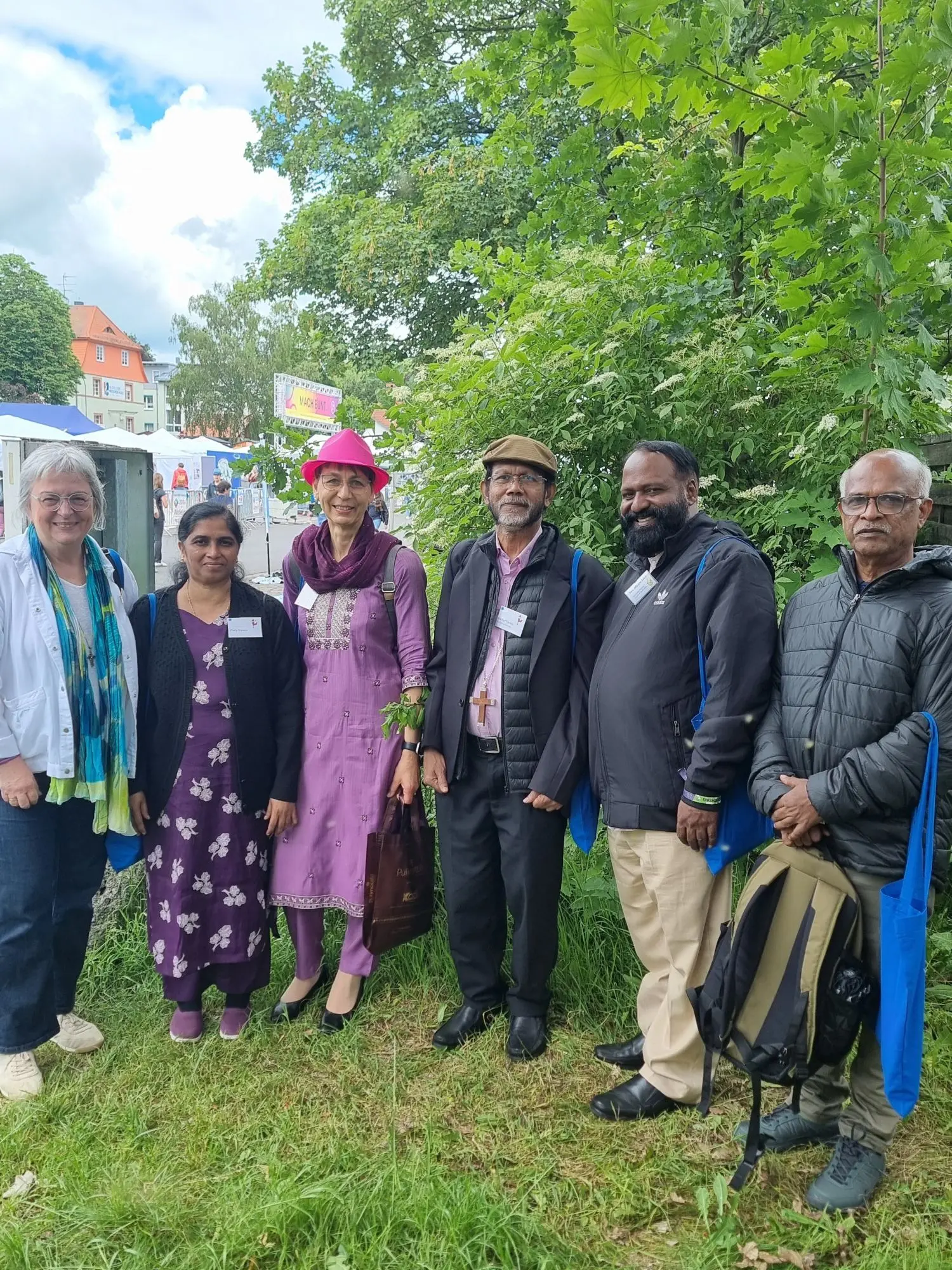 Deutsch-indische Gruppe vor einer grünen Hecke, im Hintergrund Zelte, Frauen zum Teil im Sari