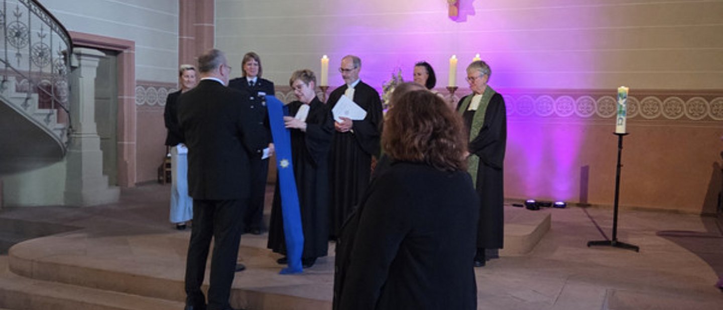 Mehrere Personen stehen vor dem Altar. Die Pfarrerin übergibt eine blaue Stola an den Oberkirchenrat, 