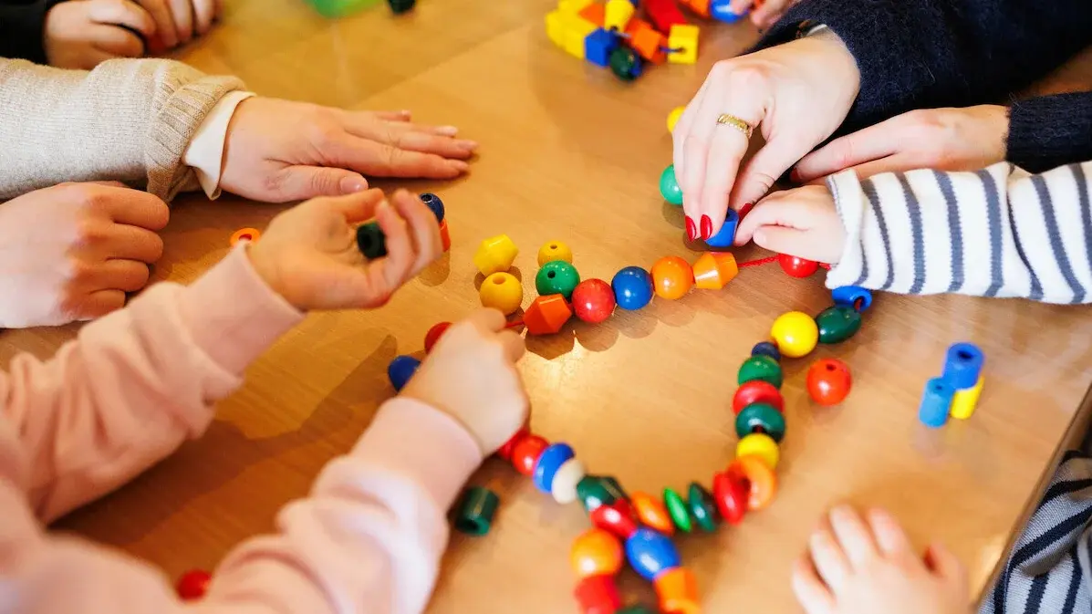 Kita - Kinder beim Spielen. An einem Tisch sitzen einige Kinder, eine Erzieherin und spielen mit einer Kette mit bunten Kugeln und anderen Formen, die auf einer Schnur aufgereiht sind.