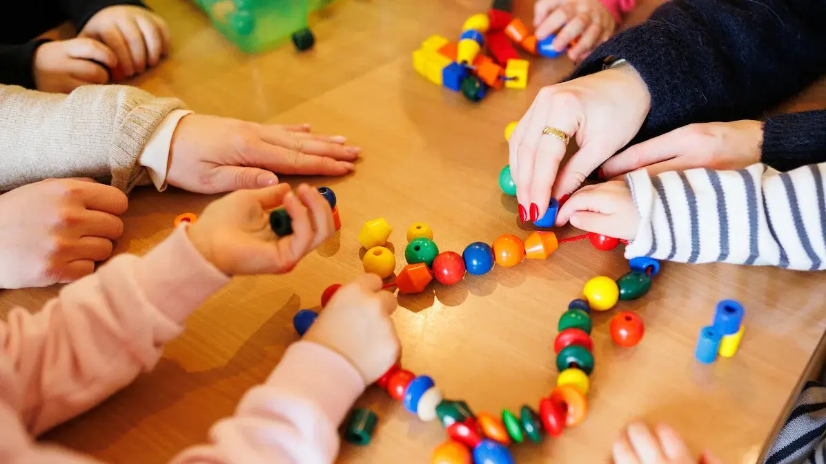 Kita - Kinder beim Spielen. An einem Tisch sitzen einige Kinder, eine Erzieherin und spielen mit einer Kette mit bunten Kugeln und anderen Formen, die auf einer Schnur aufgereiht sind.
