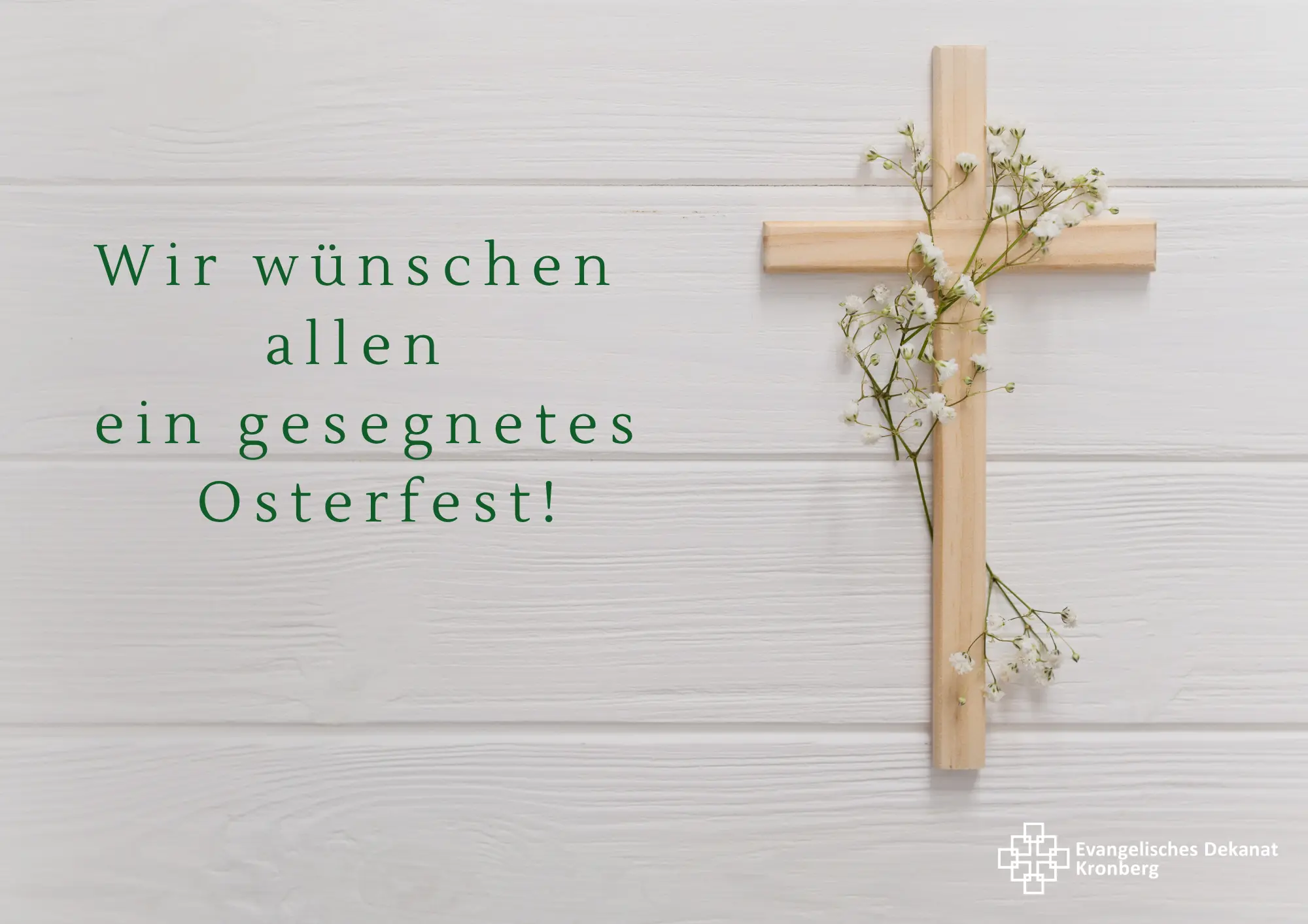 Ein mit Blumen umranktes Holzkreuz hängt an einer cremefarbenen Wand. Darauf ist der Text zu lesen: Wir wünschen  allen  ein gesegnetes  Osterfest!