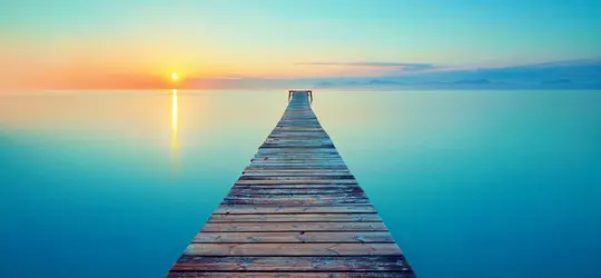 Ein langer Holzsteg führt über ruhiges Wasser in Richtung Sonnenaufgang am Horizont. Die sanften Farben des Himmels und die spiegelglatte Wasseroberfläche vermitteln Ruhe, Weite und Meditation.