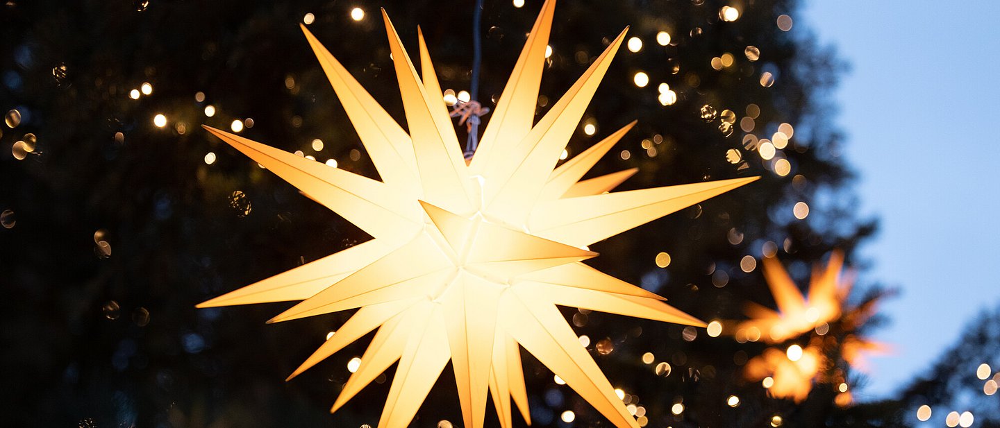 Ein Herrnhuter Stern leuchtet hell. im Hintergrund sind viele kleine funkelnde Lichter in einem Weihnachtsbaum zu erkennen.
