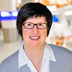Porträt einer lächelnden Frau mit kurzer dunkler Frisur und Brille. Sie trägt eine helle Bluse und befindet sich in einem hellen Flughafenumfeld mit unscharfem Hintergrund. Das Bild zeigt Frau Klünemann.
