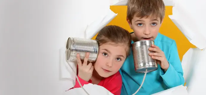 Zwei Kinder schauen durch ein Loch in einer weißen Papierwand. Sie halten ein selbstgebautes Dosentelefon: Das Mädchen hört an einer Blechdose, der Junge spricht in die andere. Beide wirken neugierig und spielerisch.