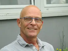 Porträt eines lächelnden Mannes mit Brille und kurzem Haar. Er trägt ein hellgraues Poloshirt und sitzt vor einer hellen Hauswand im Freien.